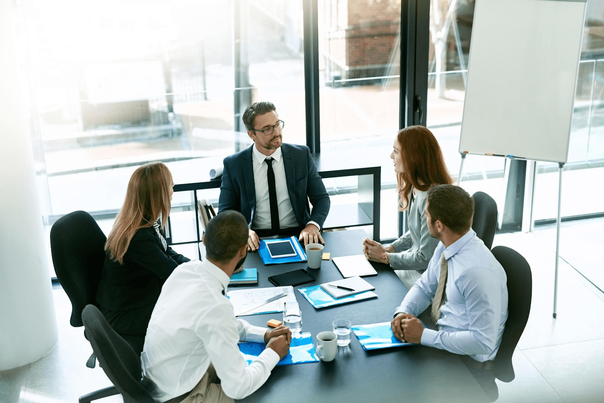 Trainer standing at a table briefing a seated team in a bright meeting room